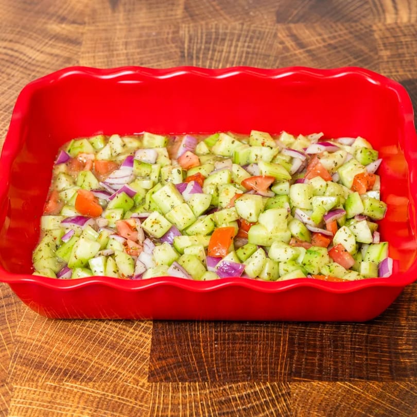 Diced cucumber, tomato, and red onion salad in a vibrant red rectangular dish on a wooden table.