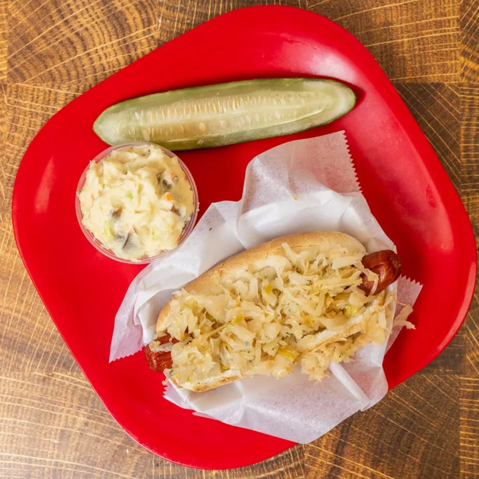 Hot dog with sauerkraut, potato salad, and a pickle spear on a red plate.