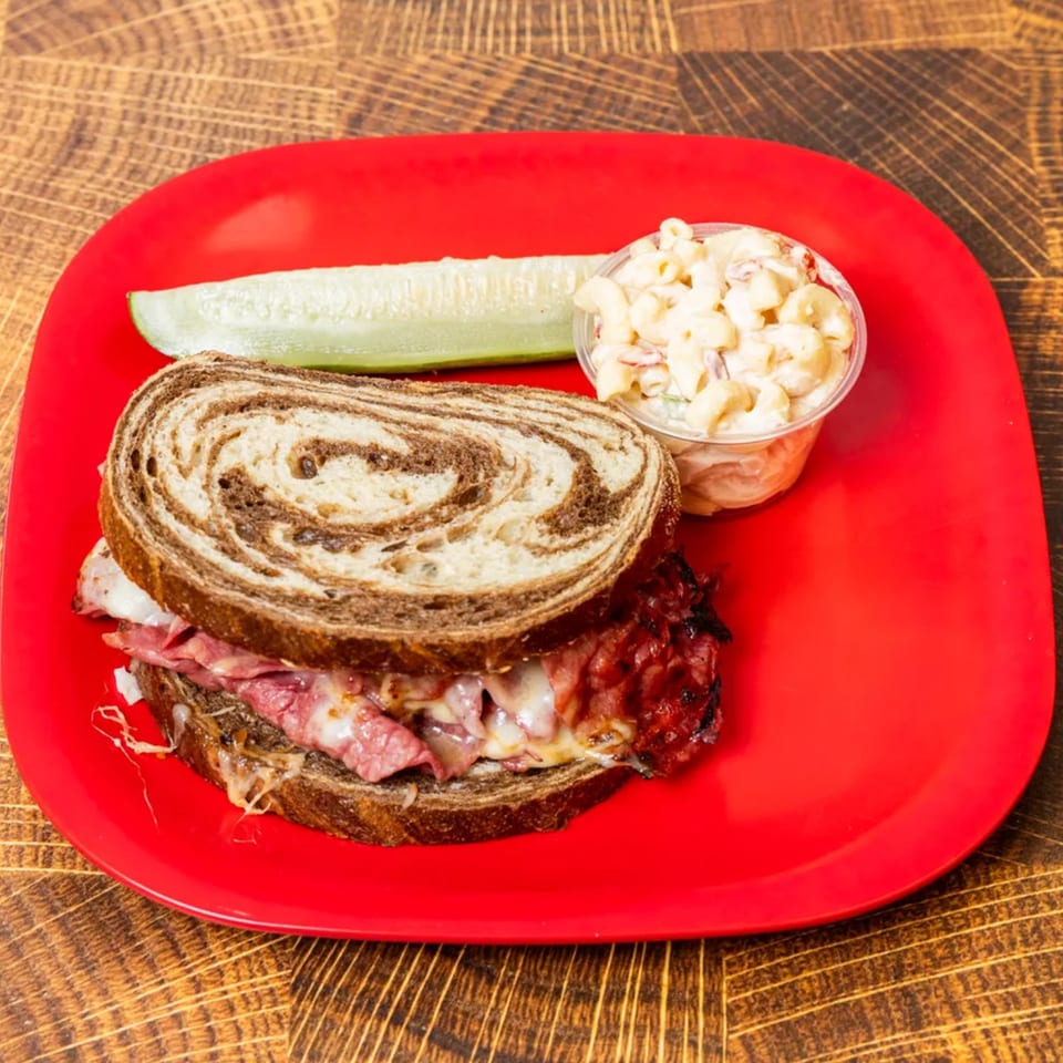 Pastrami sandwich on marble rye with melted cheese, a pickle spear, and macaroni salad on a red plate.